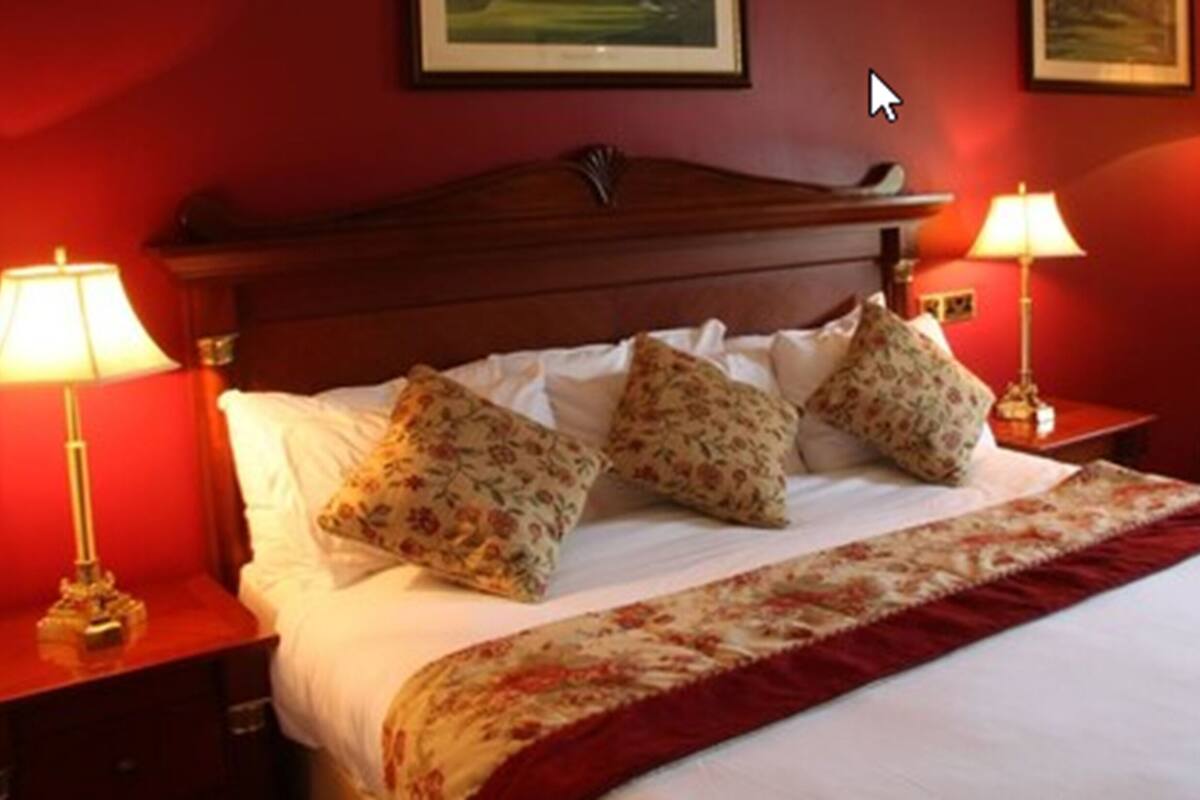 Habitación de hotel con cabecera de madera, cama grande con ropa de cama floral, dos lámparas de noche y pared roja con un cuadro enmarcado.