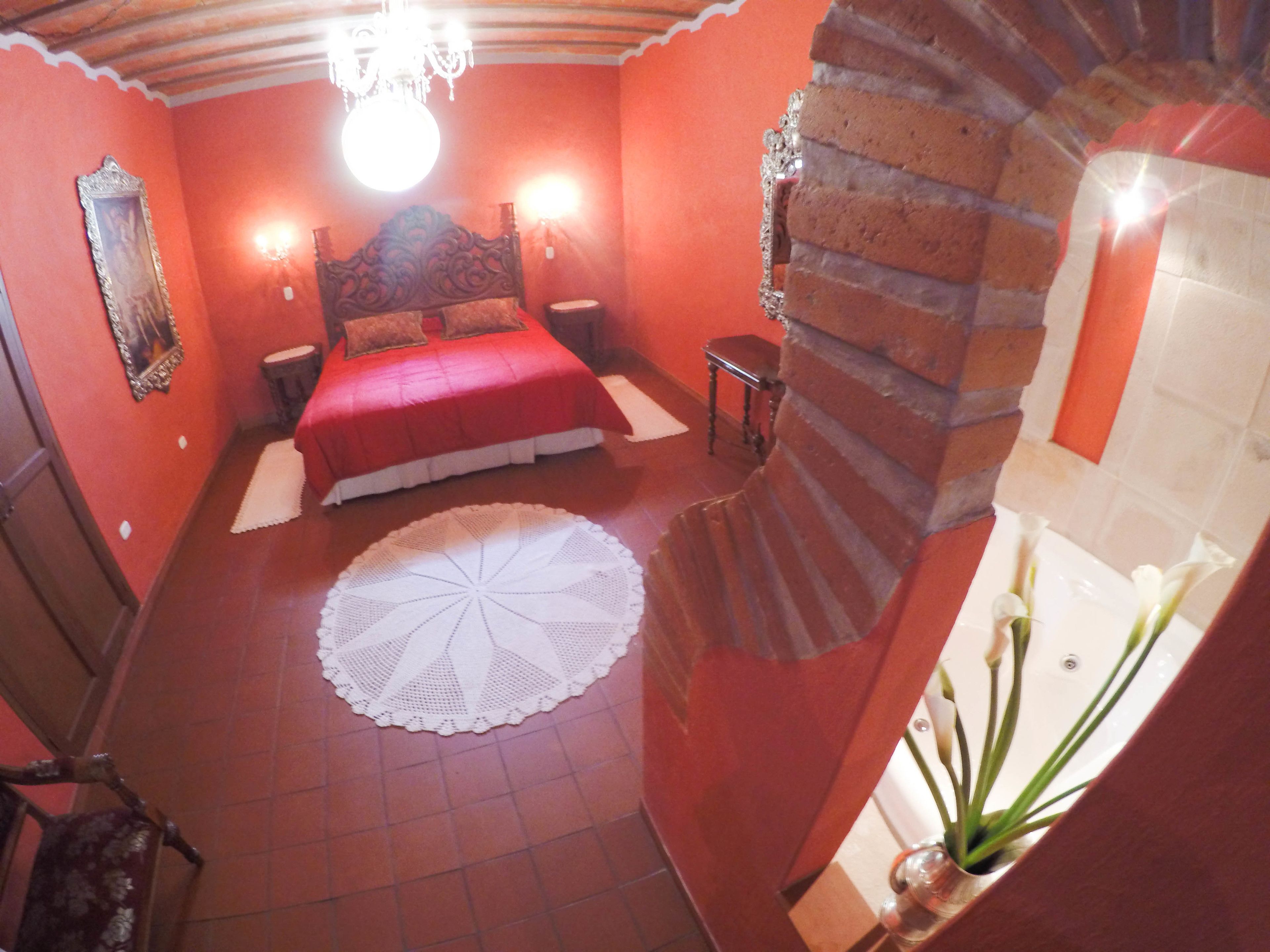 Una camera da letto con un letto rosso, un sottobicchiere rotondo, una sedia di legno, un tavolino e un punto luce fissato al muro.