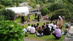 A wedding ceremony outdoors with guests seated on the grass, a tent in the background, and a scenic view of hills. - Low Wood Hall Hotel (Seascale)