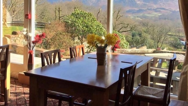 A dining area with a table and chairs, a vase with flowers, and a view of mountains through large windows. - Low Wood Hall Hotel (Seascale)