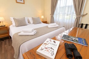 Habitación de hotel con cama bien hecha, una mesa de madera con un libro y unos binoculares, y una ventana con cortinas.