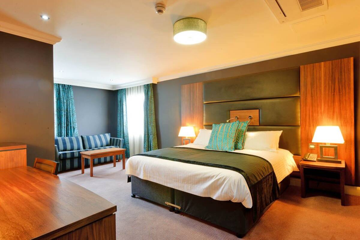 A hotel room with a large bed, two bedside tables with lamps, a wooden desk, a sofa, and a window with curtains.
