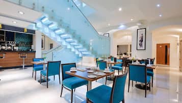 Un moderno vestíbulo de hotel con una escalera de cristal, zona de comedor con sillas azules y un bar.