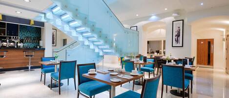 A modern hotel lobby with a glass staircase, dining area with blue chairs, and a bar.