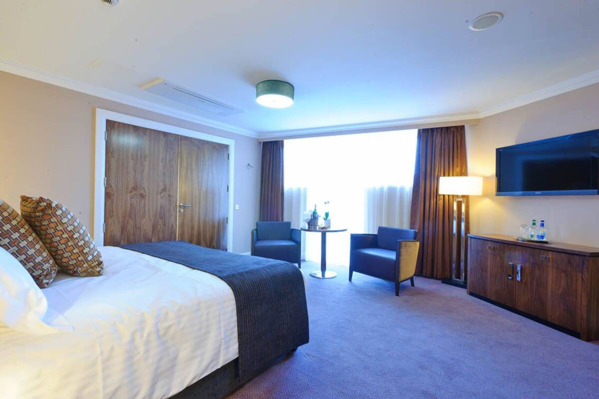 A hotel room with a large bed, two armchairs, a small table, a TV, and a wardrobe.