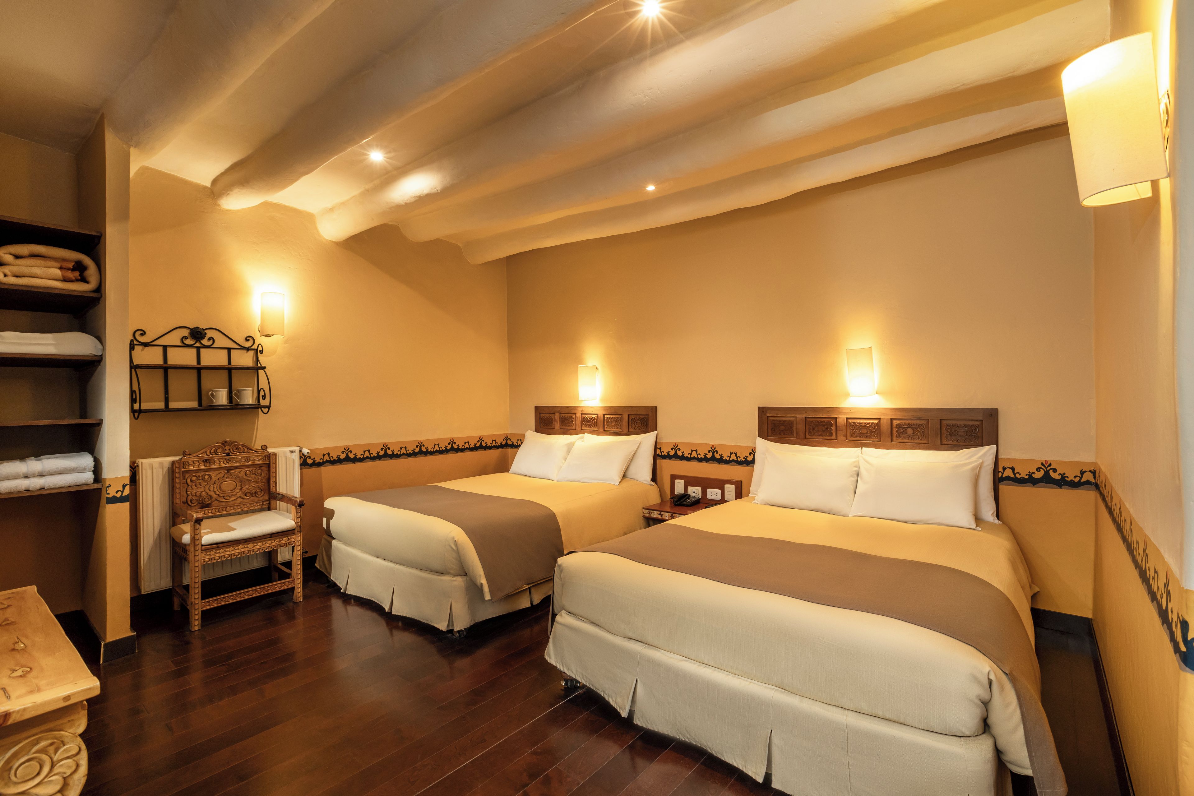 A hotel room with two beds, a wooden chair, a small table, and a shelf with towels.