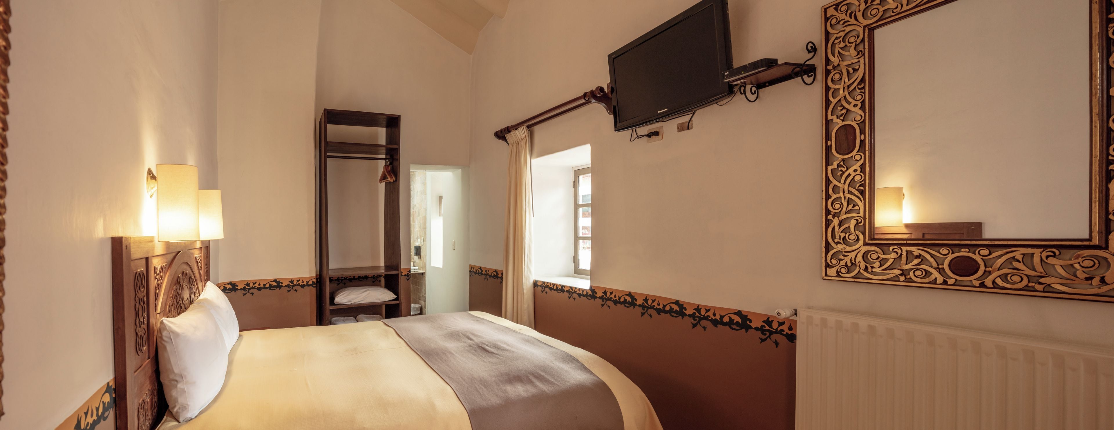 Una habitación de hotel con una cama, un televisor montado en la pared, un espejo con un marco decorativo y una ventana con cortinas.