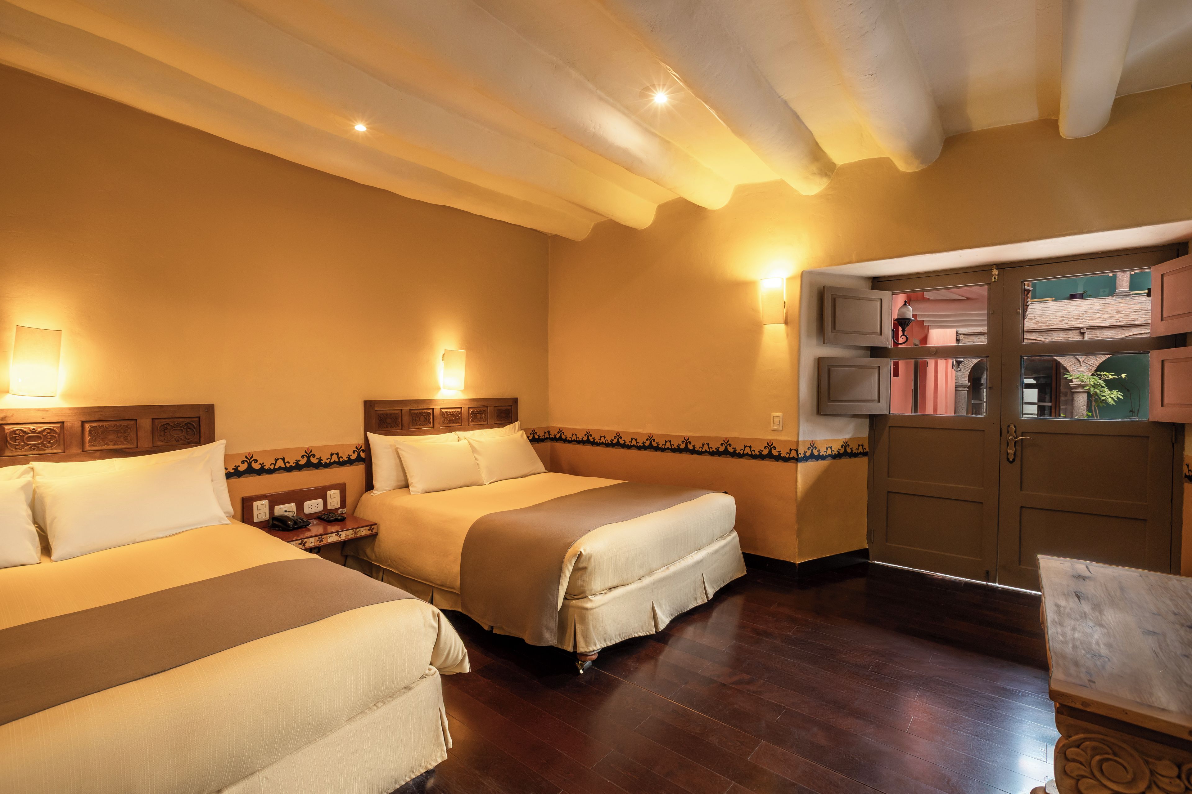 Una habitación de hotel con dos camas, cabecero de madera, mesita de noche, lámpara de pared y un armario de madera con espejo.