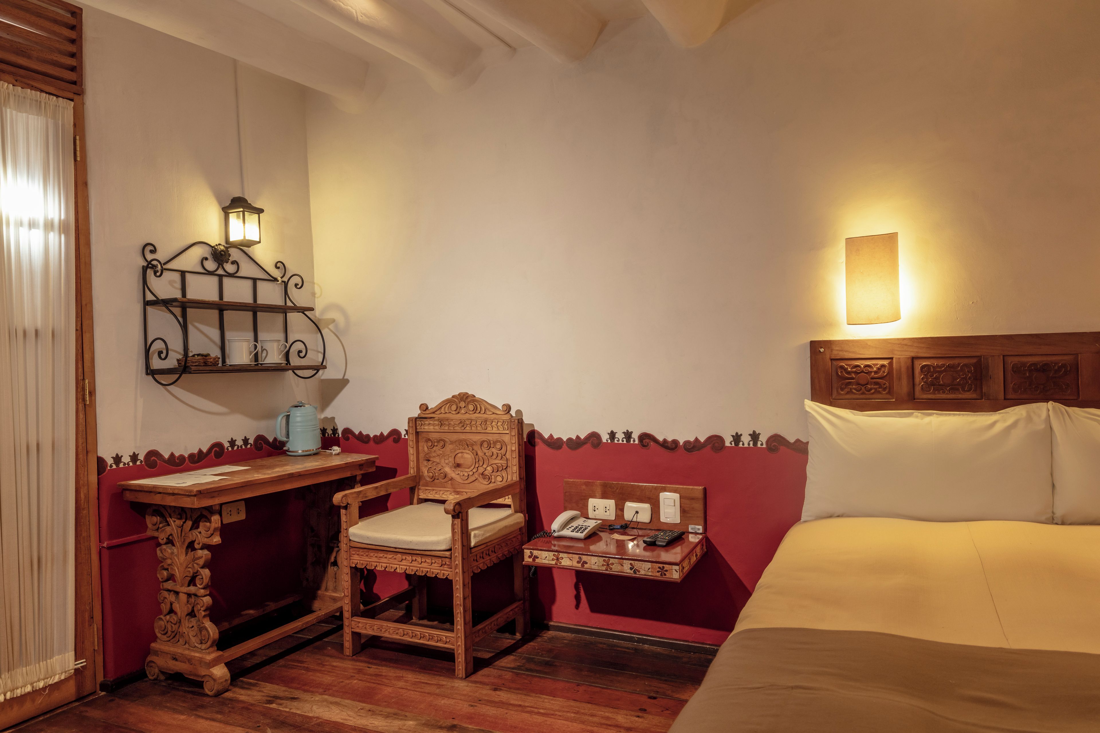 A hotel room with a wooden desk, a chair, a bedside table with a lamp, a bed, and a wall-mounted telephone.