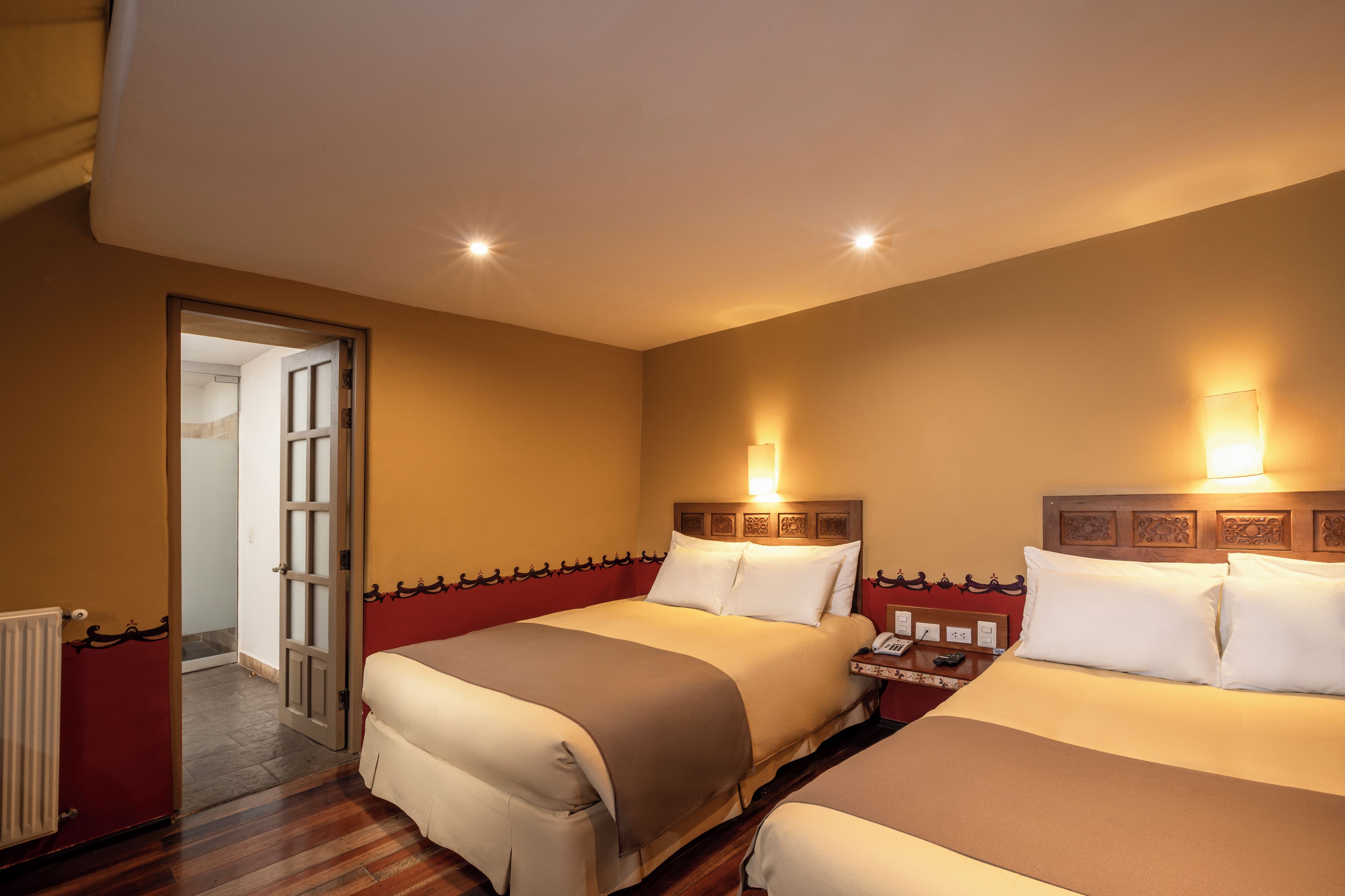 A hotel room with two beds, a wooden headboard, bedside lamps, a nightstand, and a door leading to another room.
