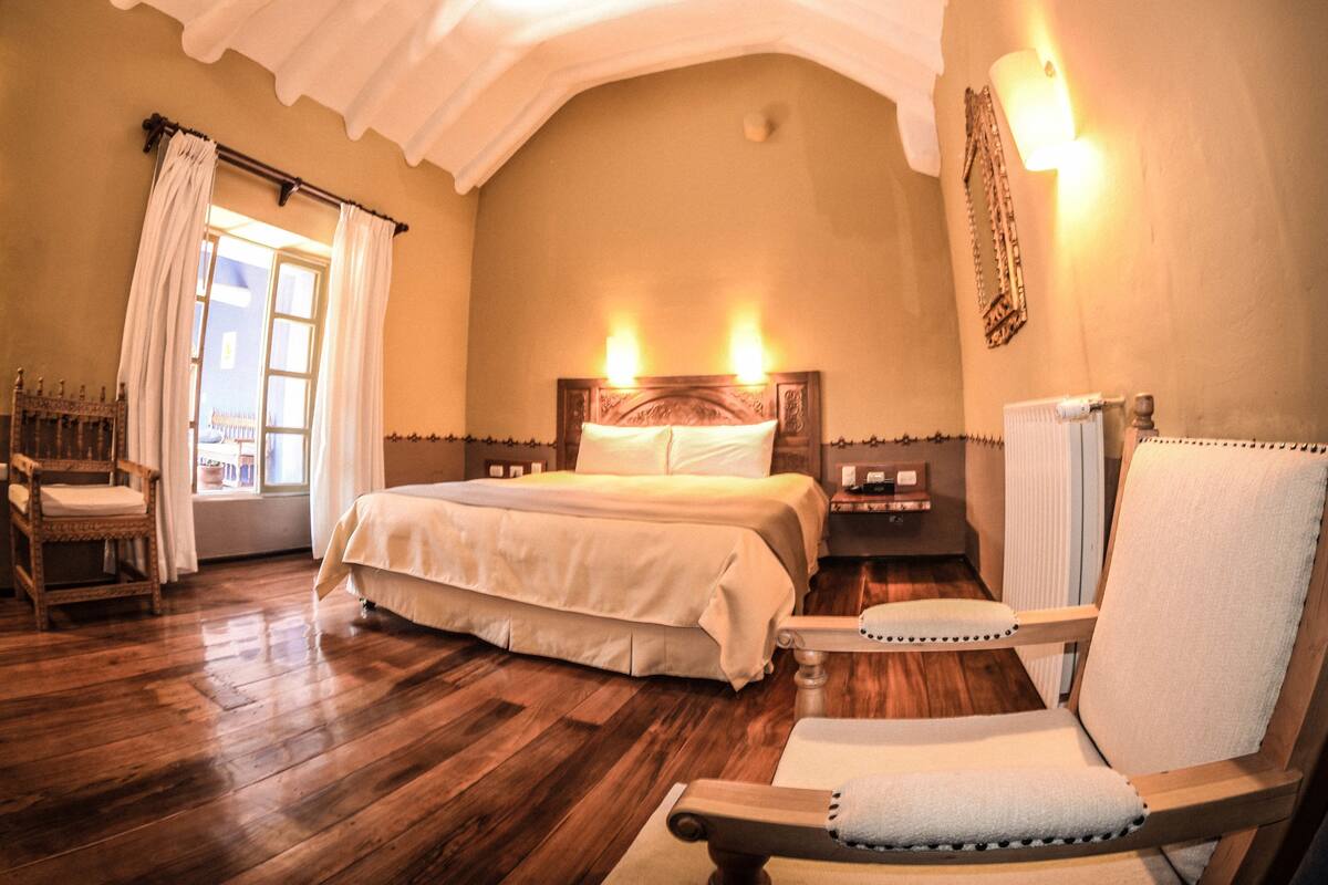 Ein Schlafzimmer mit einem großen Bett, einem Holzsessel, einem Nachttisch und einem Fenster mit Vorhängen.