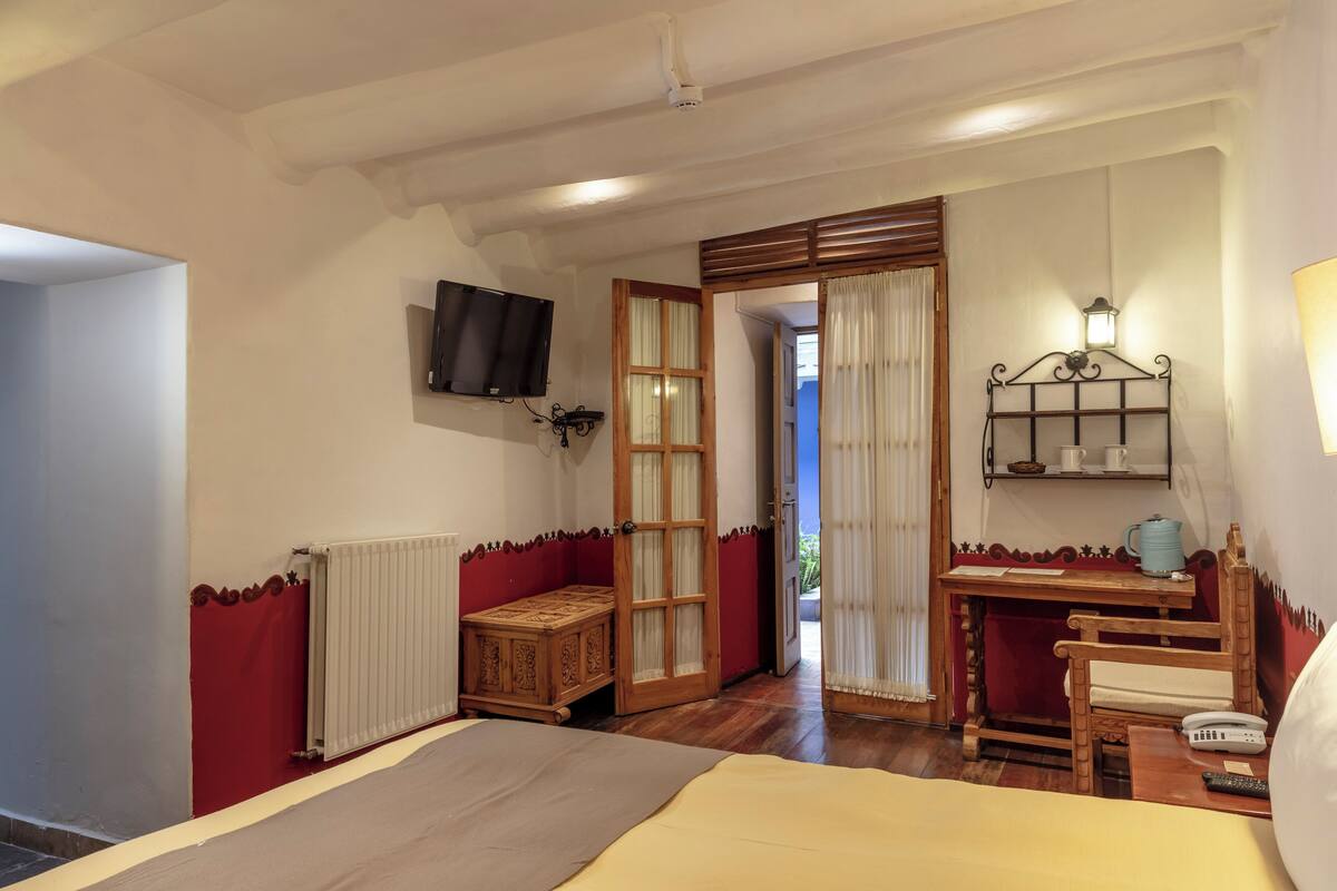 Ein Zimmer mit einem Bett, einer Holzkiste, einem Tisch mit einer Lampe und einem an der Wand montierten Fernseher.