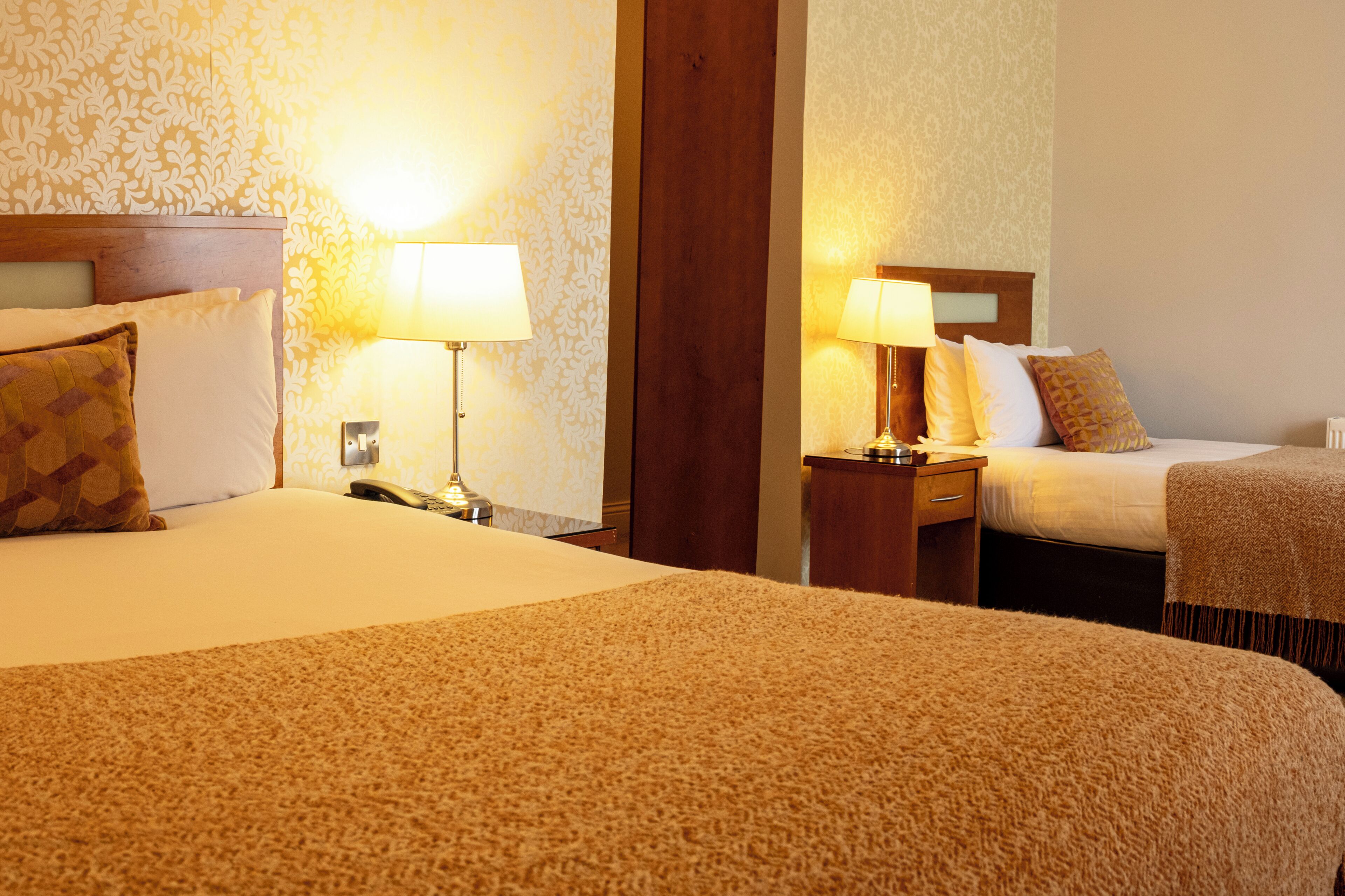 A hotel room with two beds, each with a wooden headboard, bedside tables, lamps, and patterned wall wallpaper.