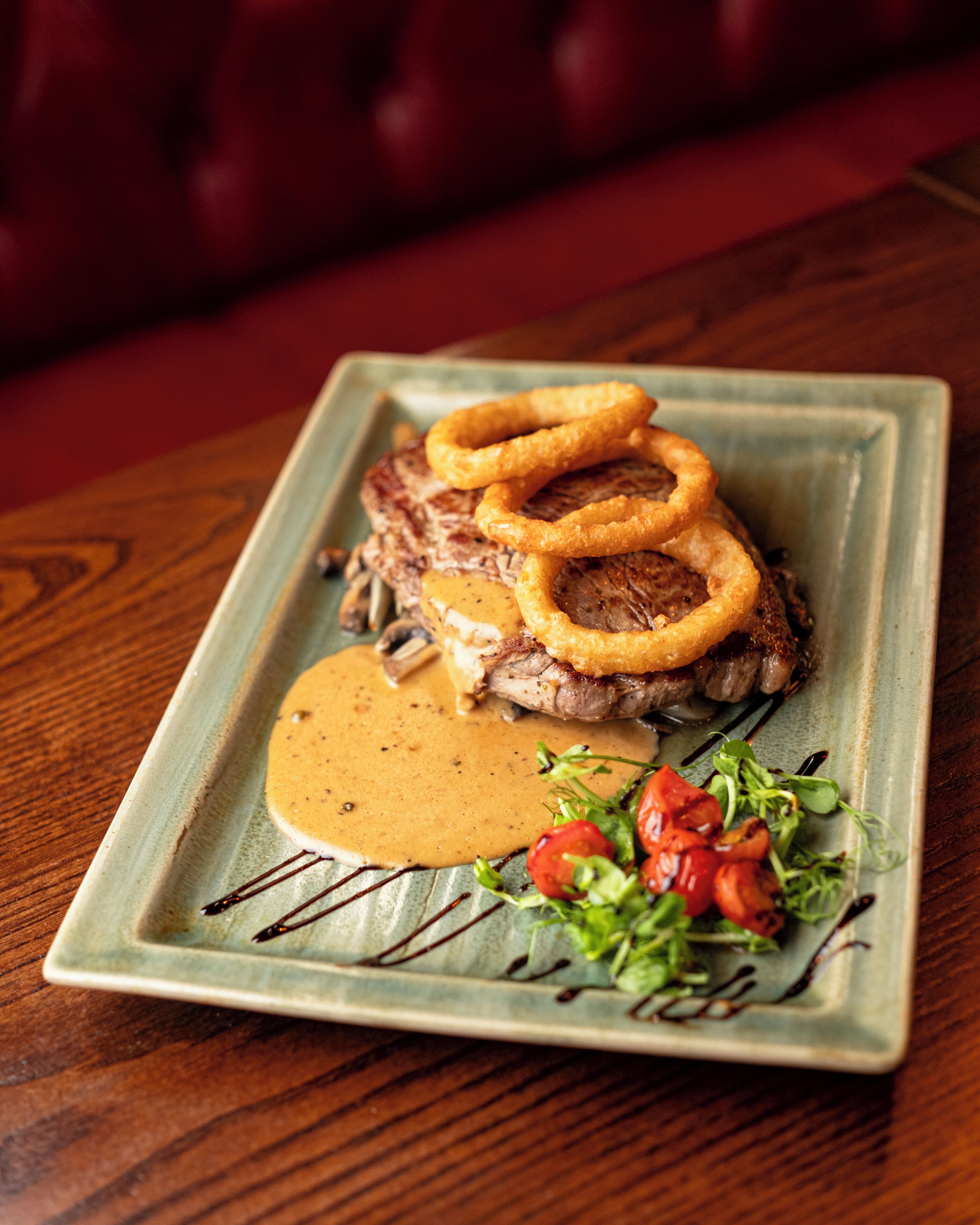 A square plate with a steak, onion rings, a creamy sauce, and a side of greens.