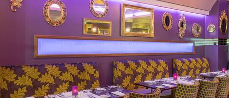 A restaurant with purple walls, ornate mirrors, and patterned upholstery. Tables are set with white tablecloths and yellow chairs.