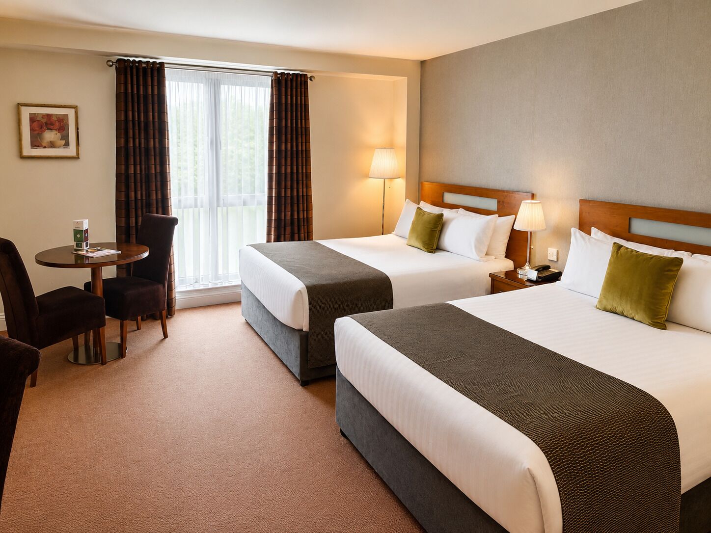 A hotel room with two beds, a desk, chairs, a lamp, and a window with curtains.
