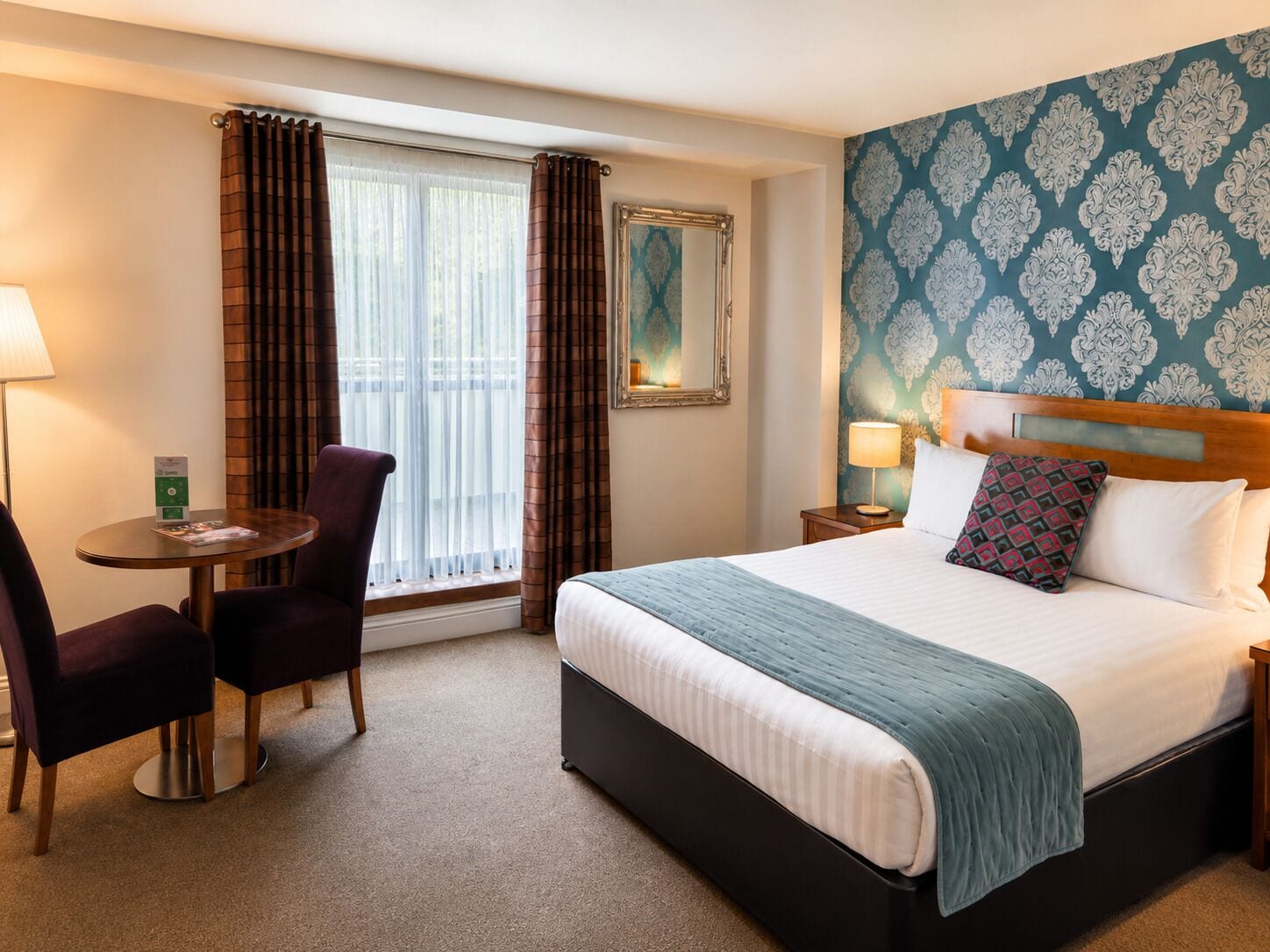 A hotel room with a bed, a desk with a chair, a window with curtains, a nightstand with a lamp, and a wall with a patterned wallpaper.