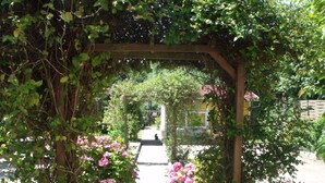 Ein Gartenweg unter einem hölzernen Bogen, der mit Kletterpflanzen und Blumen bewachsen ist.