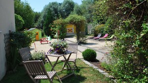 Eine Terrasse mit Tisch und Stühlen, ein kleines gelbes Gebäude und ein Garten mit verschiedenen Pflanzen und einer Bank.