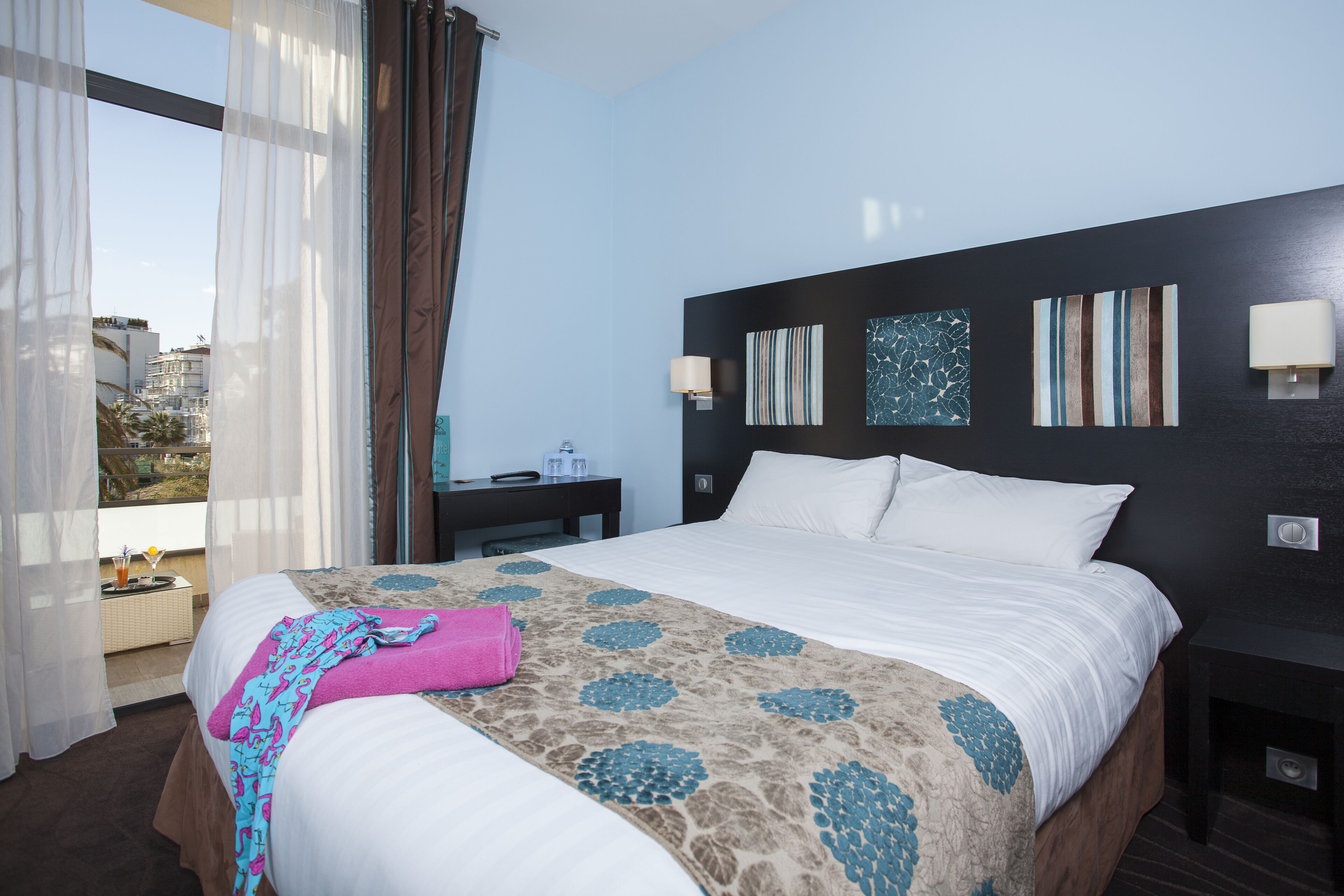 A hotel room with a large bed, a nightstand, a window with curtains, and artwork on the wall.