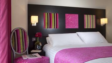 Habitación de hotel con cama, mesita de noche con lámpara, tocador con espejo y una obra de arte en la pared.