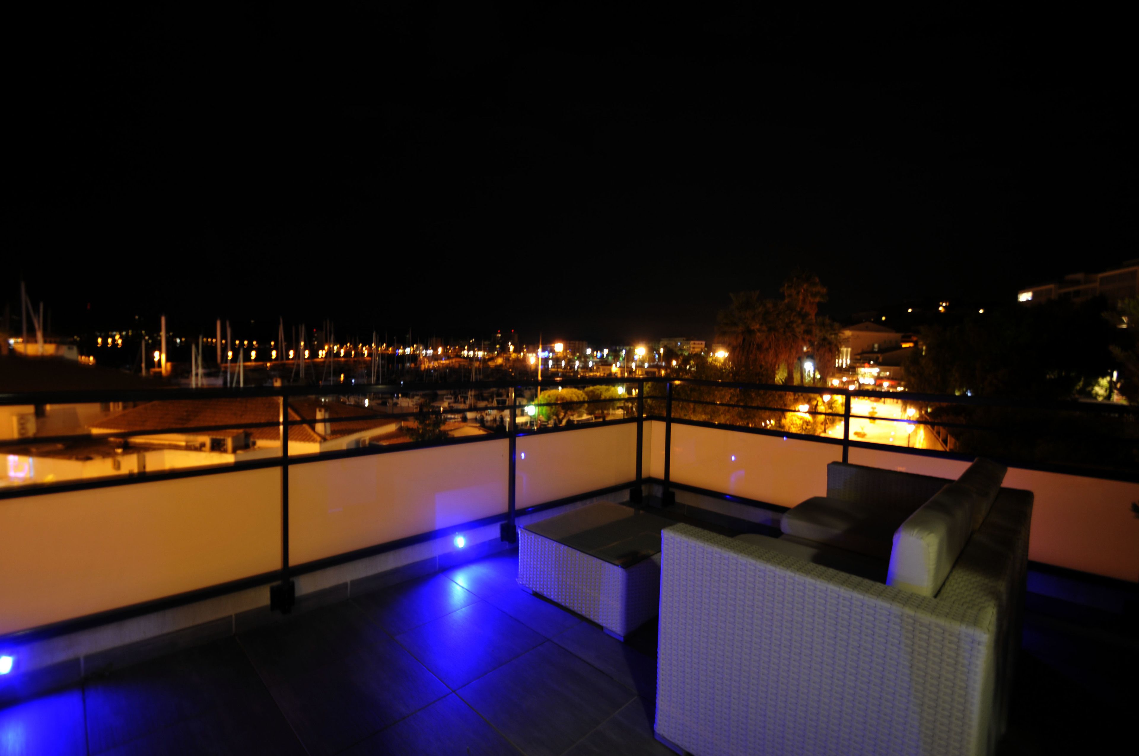 Een terras op het dak met uitzicht op een jachthaven bij nacht, met een zithoek met kussens en een balustrade.
