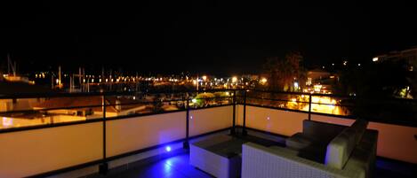 Een terras op het dak met uitzicht op een jachthaven bij nacht, met een zithoek met kussens en een balustrade.
