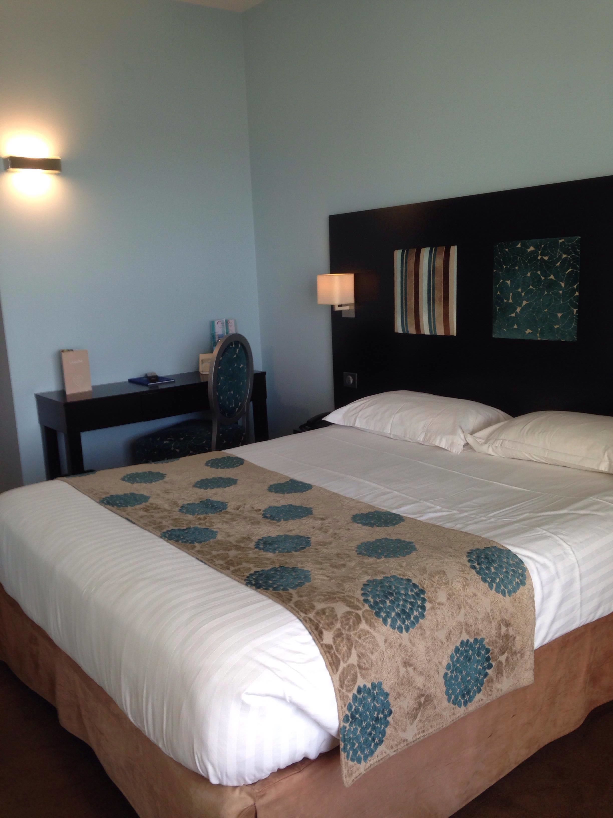 A hotel room with a large bed, a desk, and a chair. There are two wall-mounted lamps and a painting on the wall.