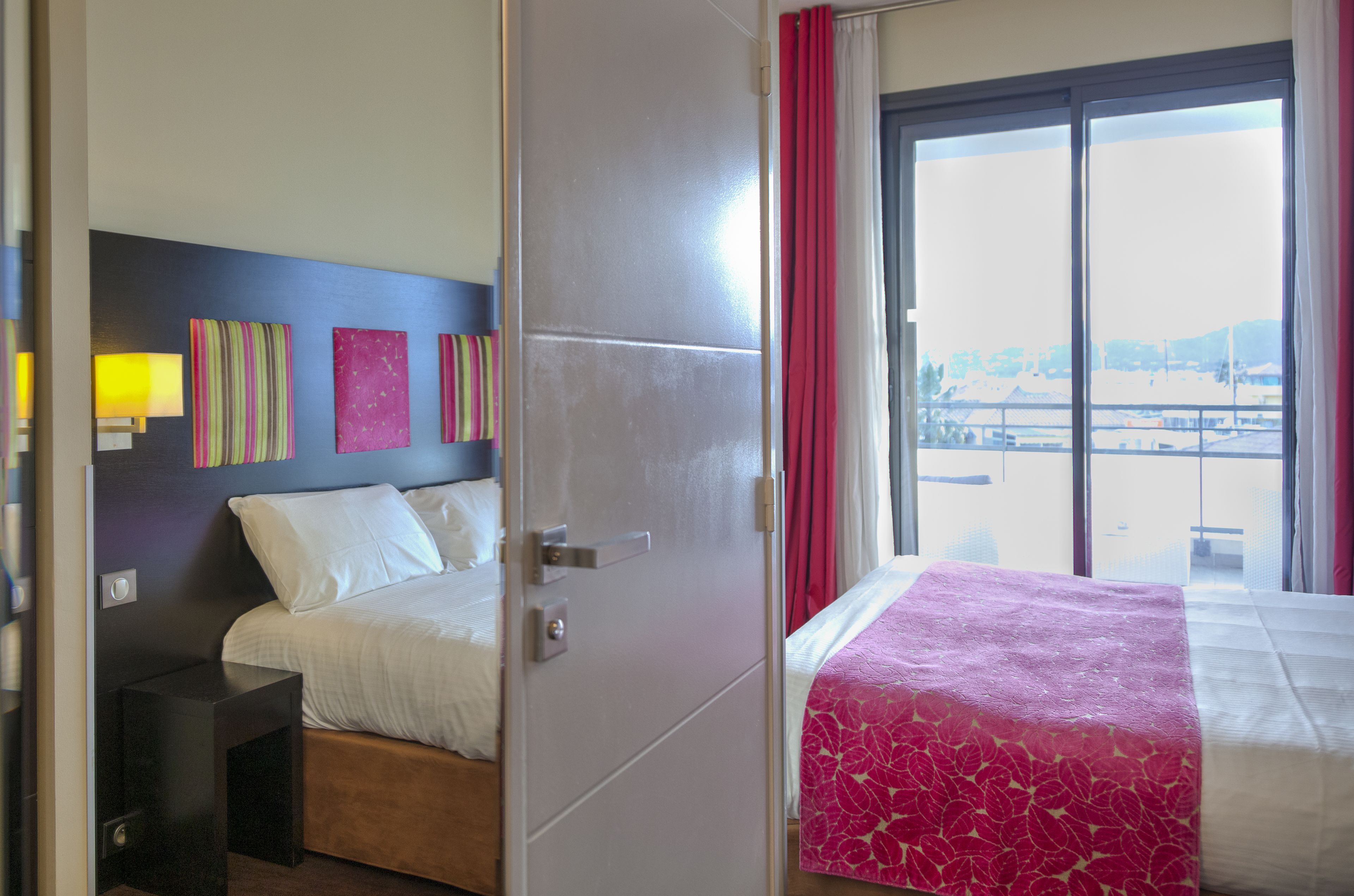 A hotel room with a bed, a nightstand, a window with red curtains, and artwork on the wall.