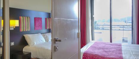 A hotel room with a bed, a nightstand, a window with red curtains, and artwork on the wall.