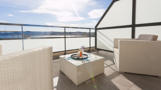 Een balkon met rieten meubilair, een glazen tafel met een drankje, en uitzicht op een waterpartij en verre gebouwen.