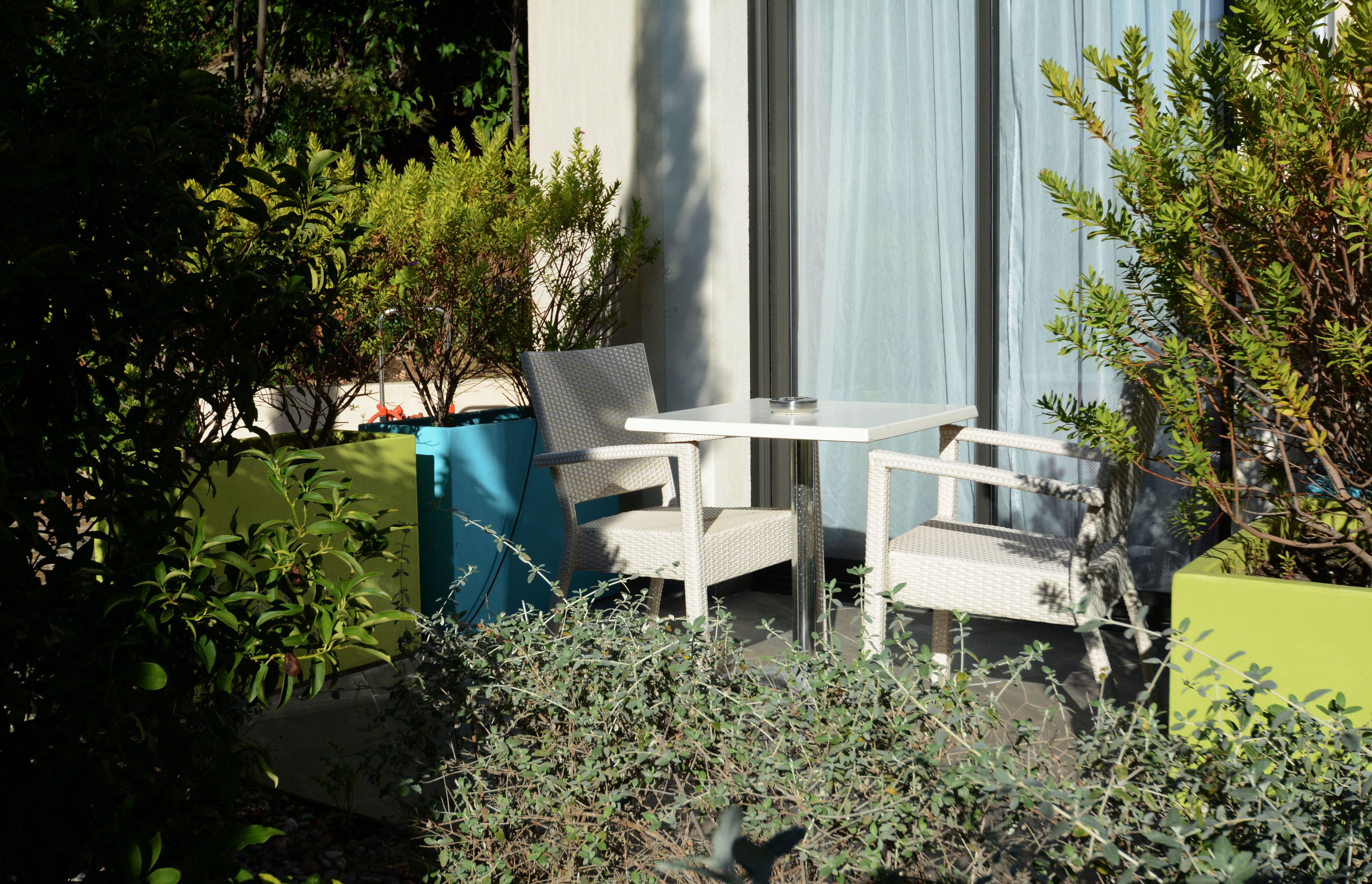 Une terrasse avec une table blanche et deux chaises, entourée de verdure et bordée d’un mur bleu.