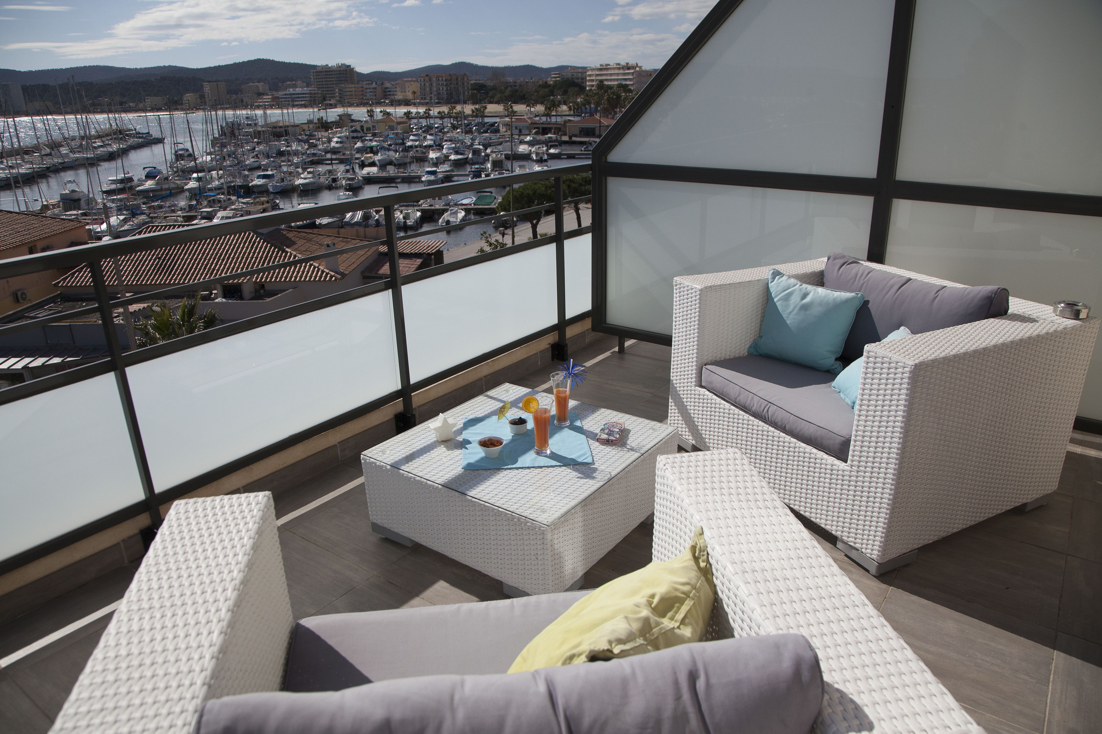 A balcony with white wicker furniture, a table with drinks, and a view of a marina and buildings.
