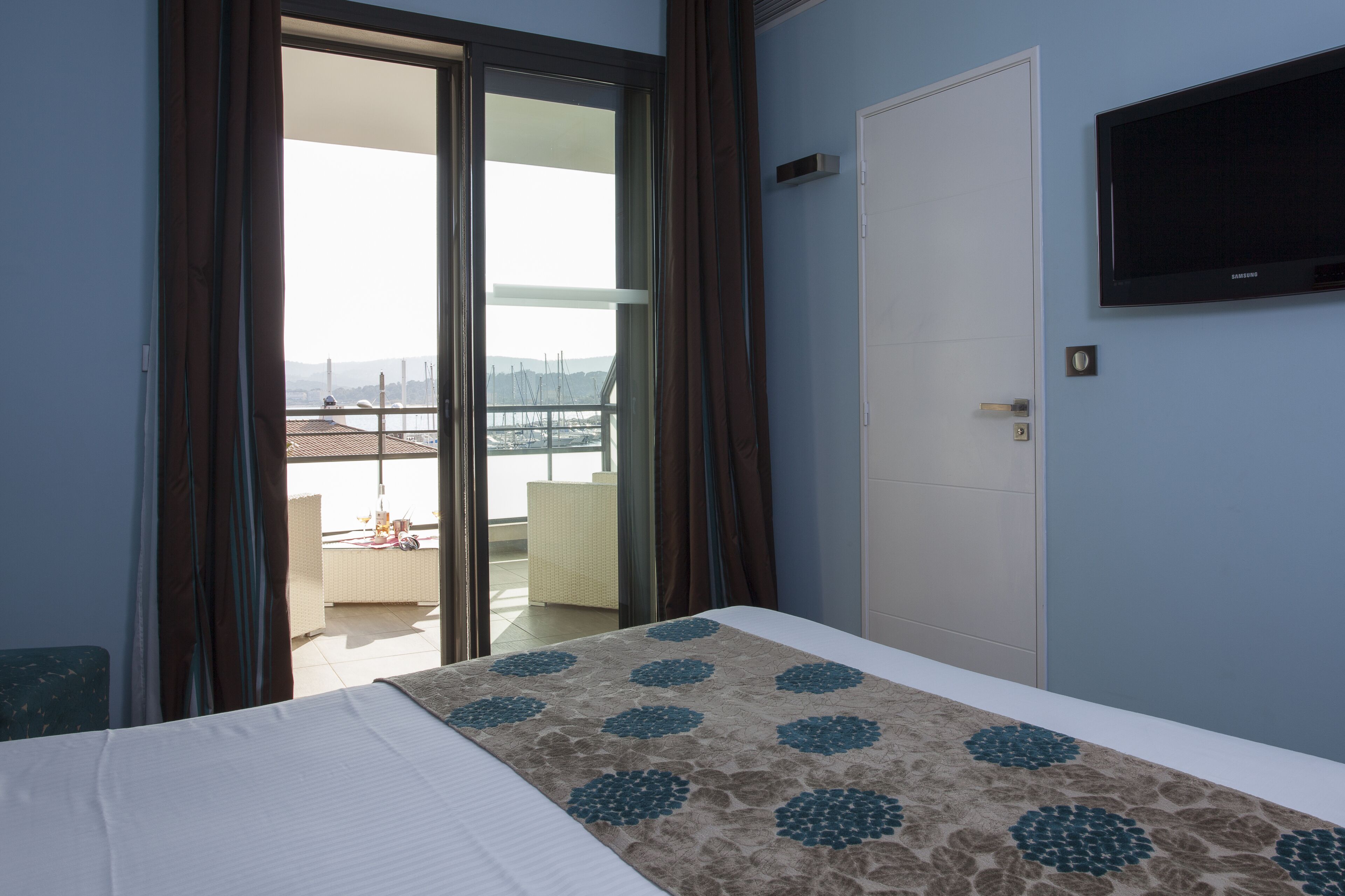 A hotel room with a bed, a TV, a door, and a view of the outside balcony.