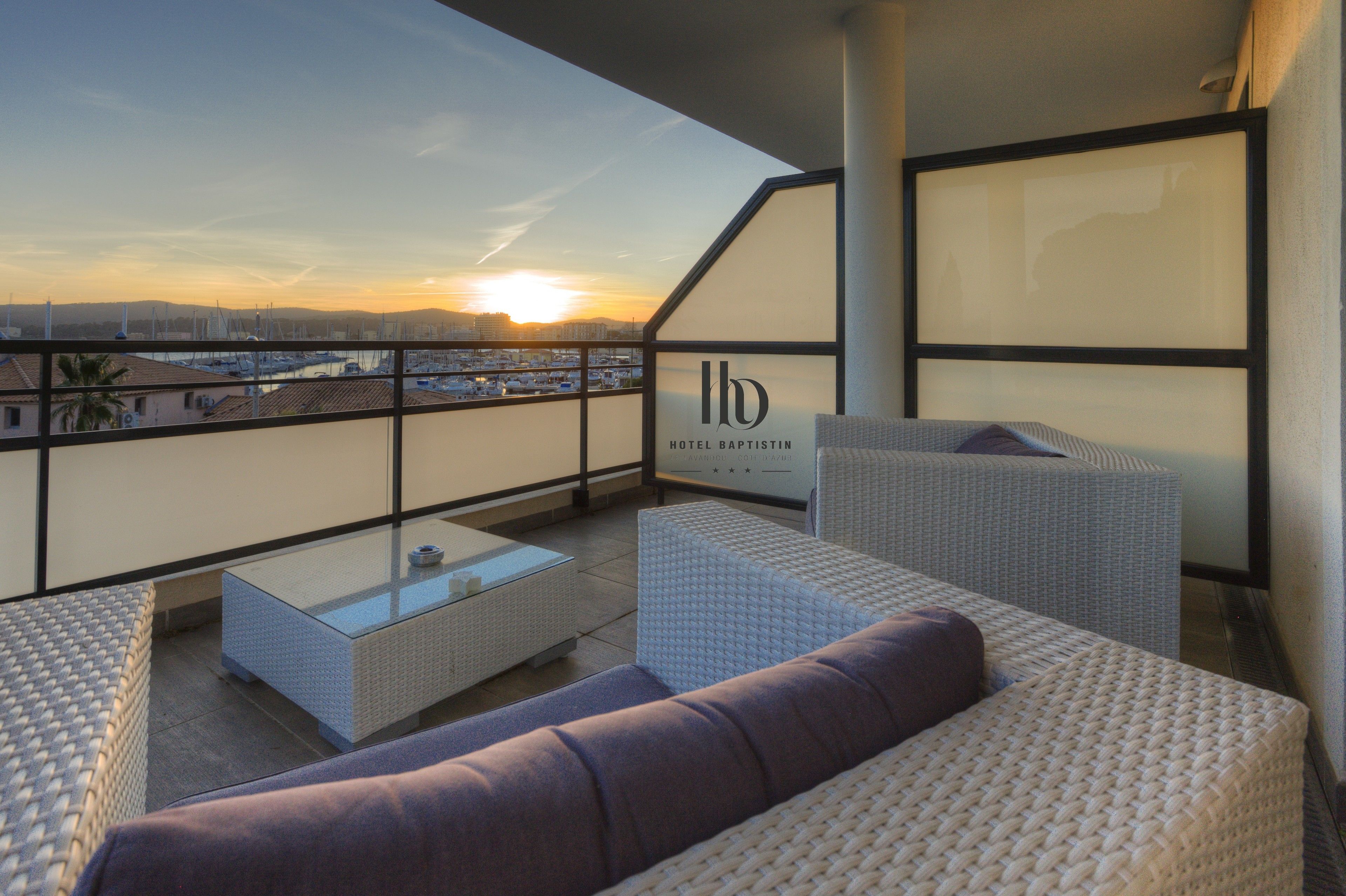 Een balkon met rieten meubels, een glazen bijzettafeltje en uitzicht op een jachthaven in de zonsondergang.