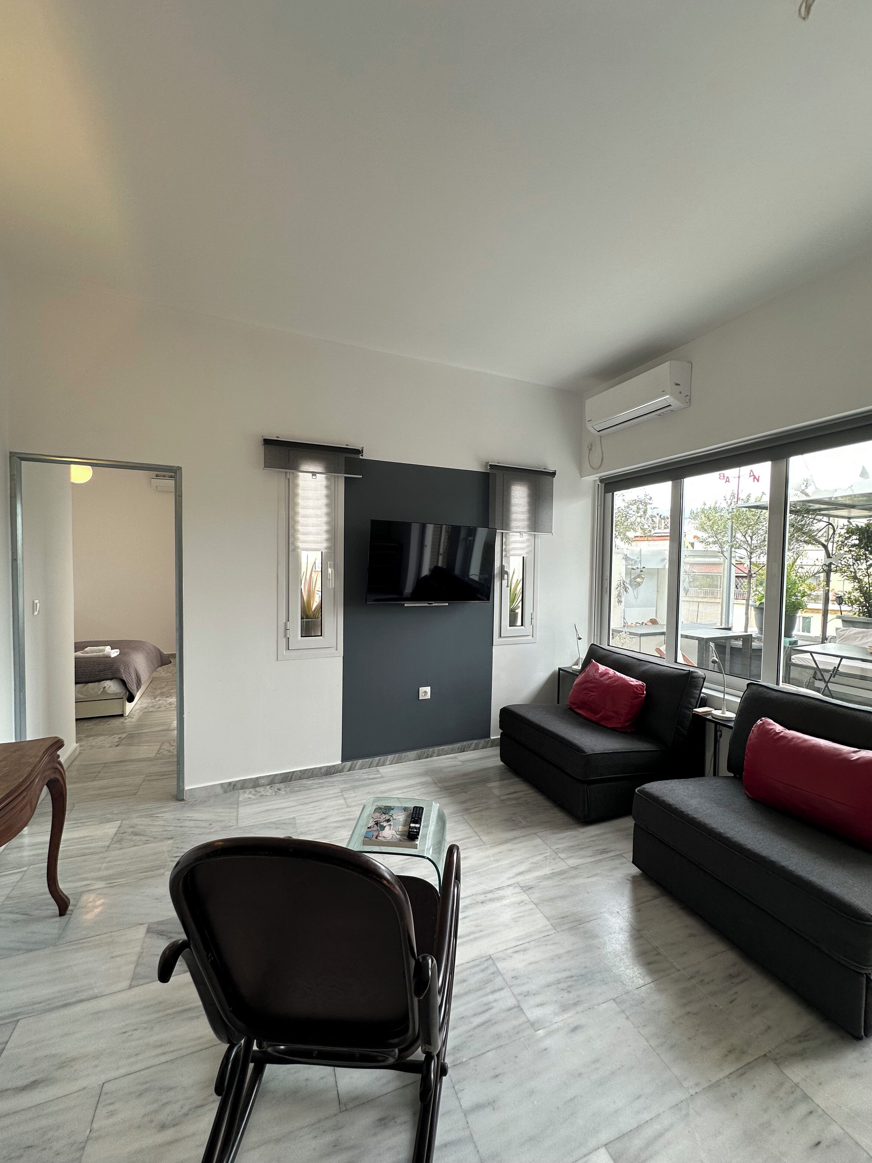 Een moderne woonkamer met een bank, een fauteuil, een aan de muur bevestigde televisie en een groot raam.