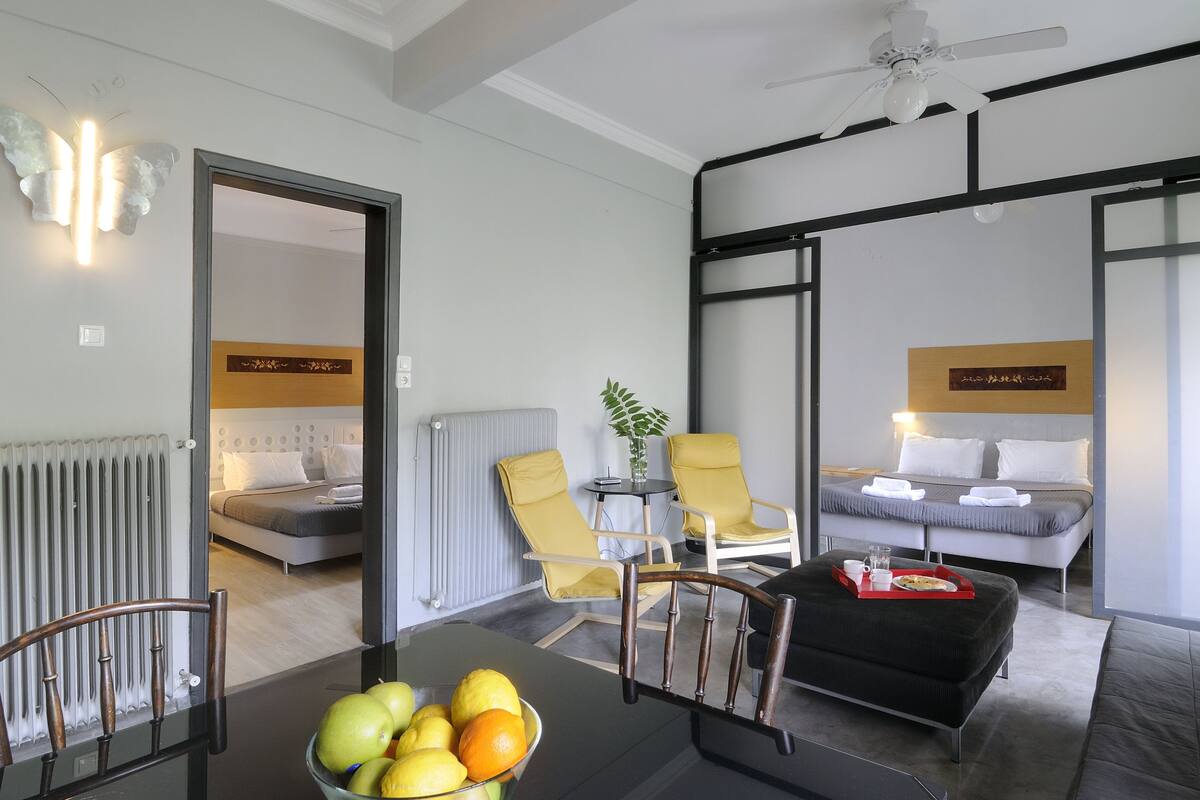 A modern hotel room with a bed, two yellow chairs, a table with fruit, and a ceiling fan.