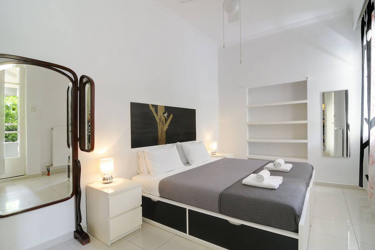 A modern bedroom with a large bed, a bedside table with a lamp, a mirror, and a painting on the wall.