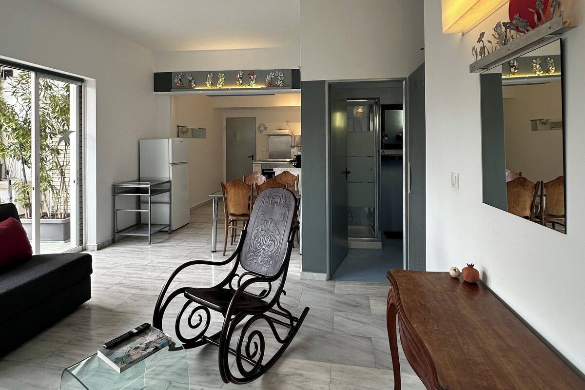 A modern living room with a glass coffee table, a wooden side table, a rocking chair, and a dining area in the background.
