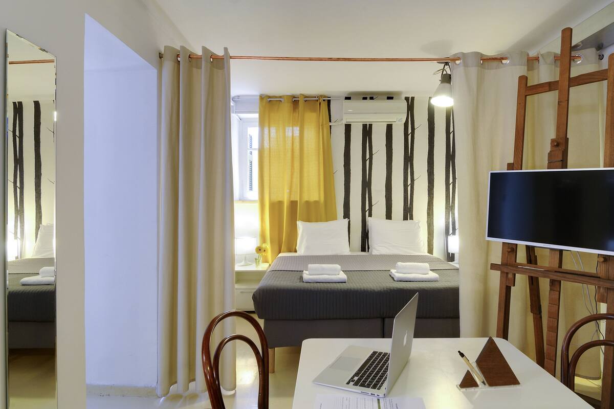 A hotel room with a bed, a desk with a laptop, a TV, and a window with curtains.