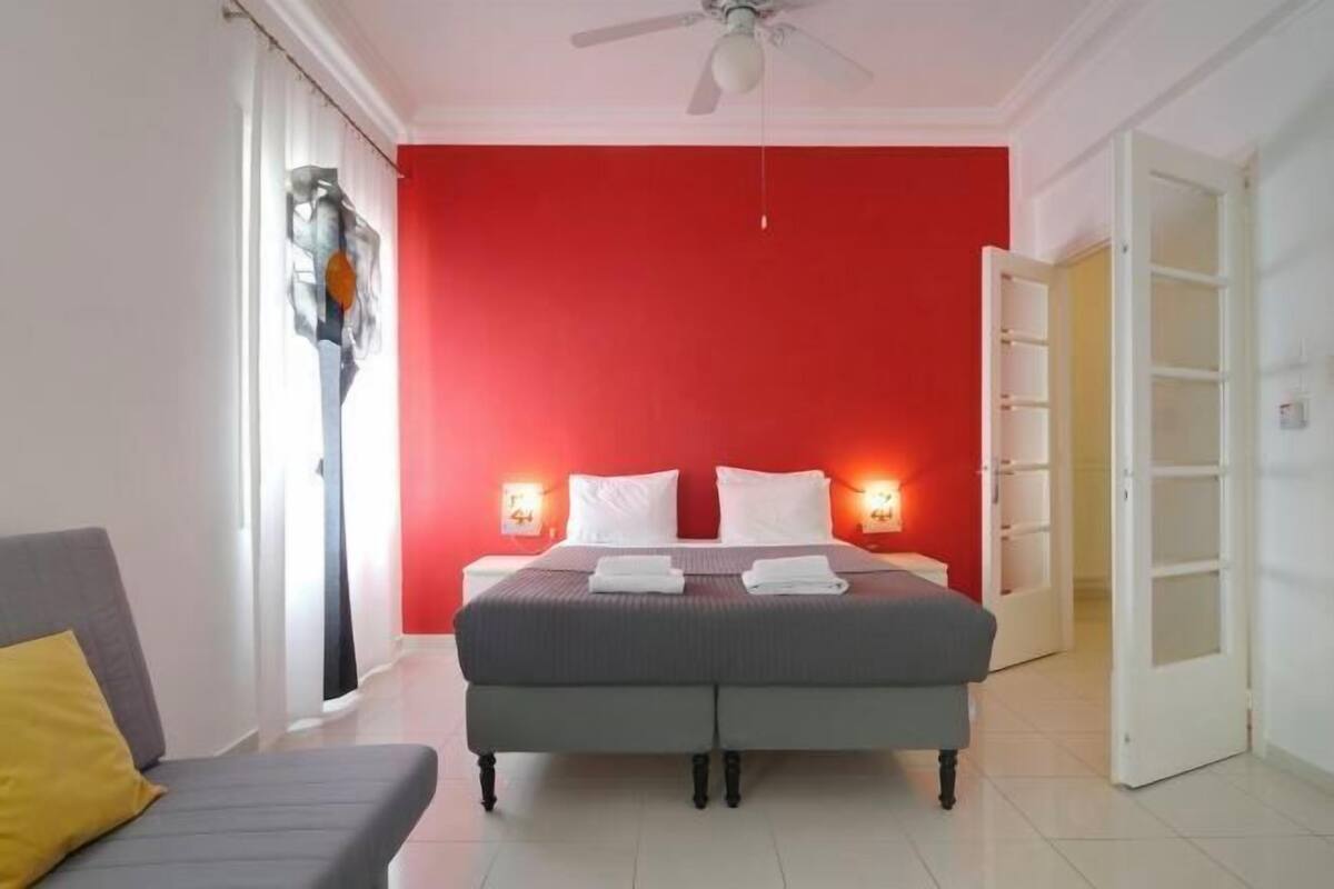 A bedroom with a red wall, a bed with white linens, a grey sofa, and a ceiling fan.