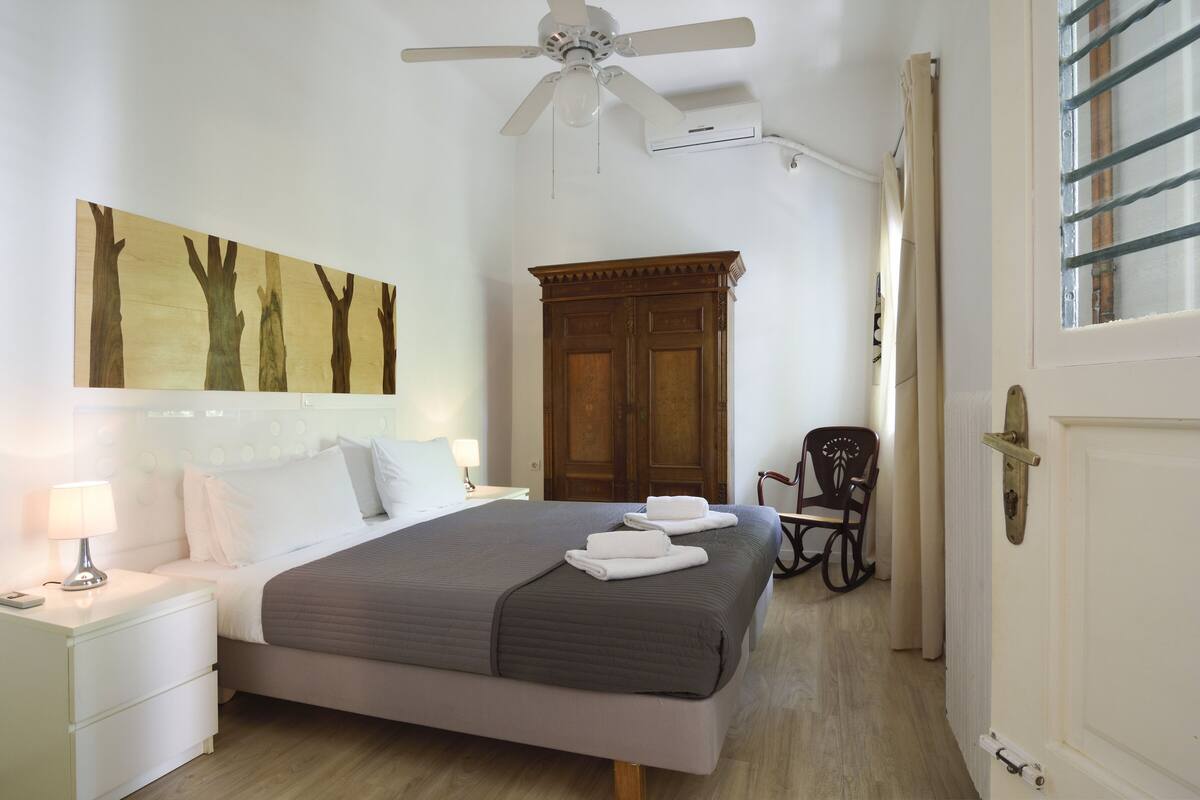 A bedroom with a bed, nightstand, wall art, ceiling fan, wardrobe, and a chair.