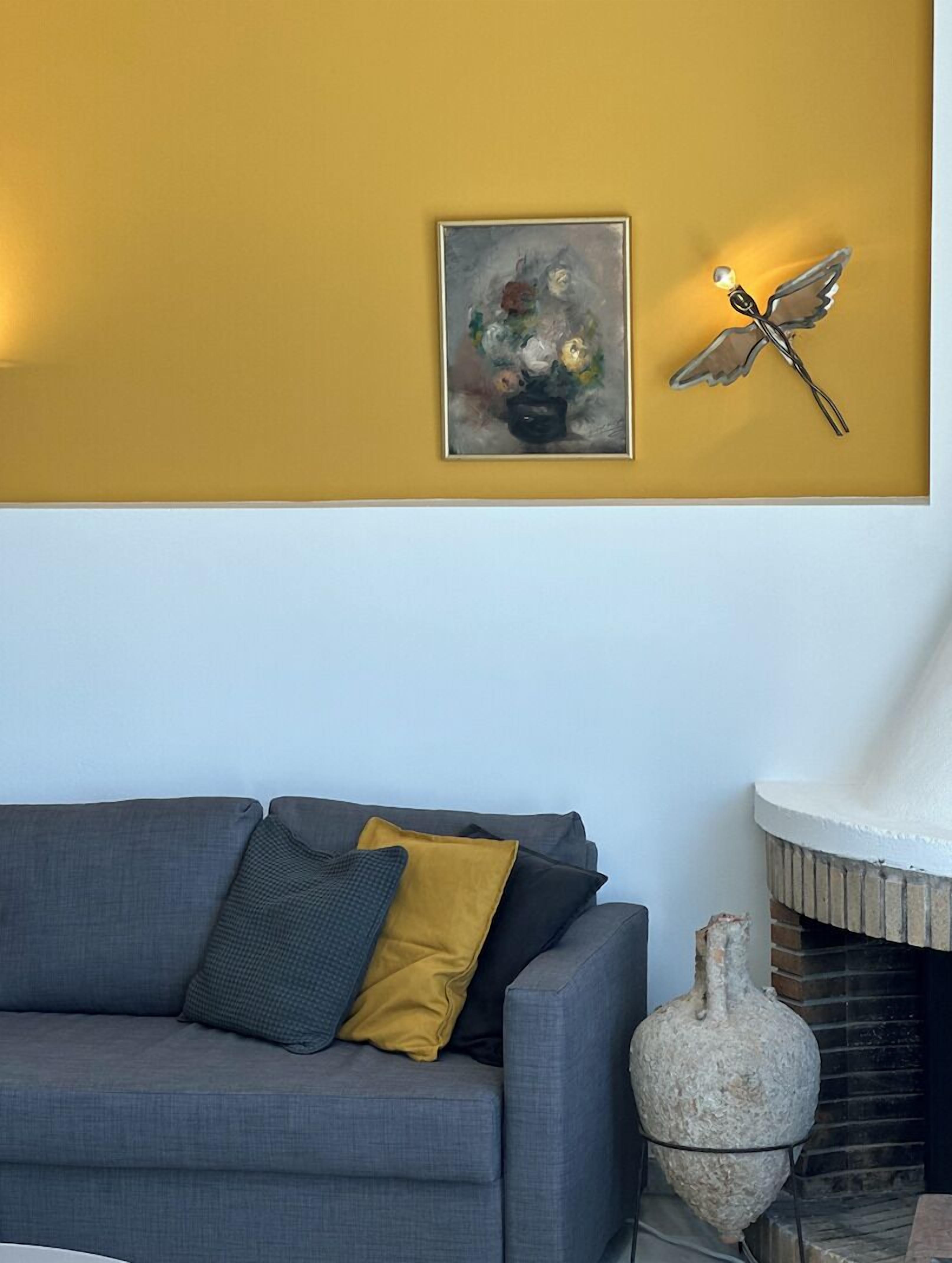 Un salon avec un canapé gris, un coussin jaune, un tableau et un luminaire fixé au mur.