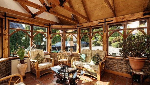 A sunroom with wooden beams, large windows, wicker furniture, and a chess table. - Tudor Lodge (Glendalough)