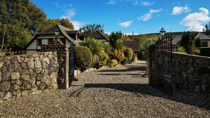 A gravel driveway leading to a stone wall and a gated entrance to a property with a house and a lamp post. - Tudor Lodge (Glendalough)