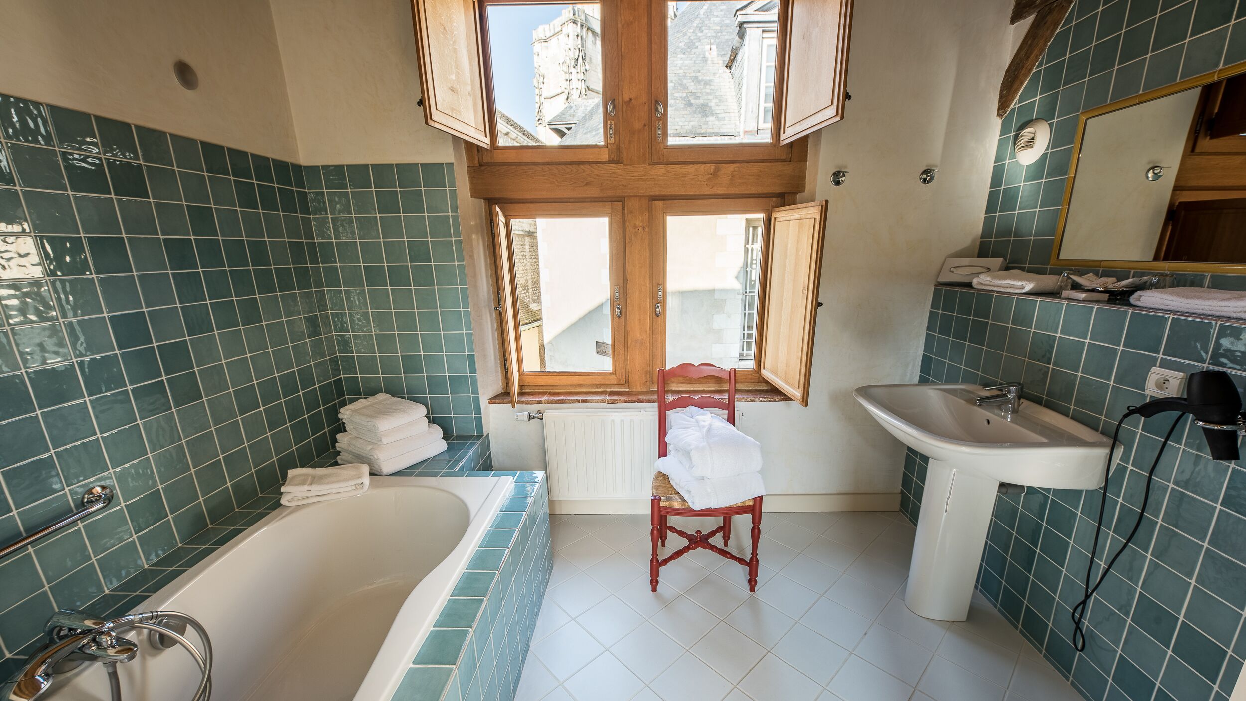 Un baño con bañera, lavabo, una silla y una ventana con contraventanas de madera.