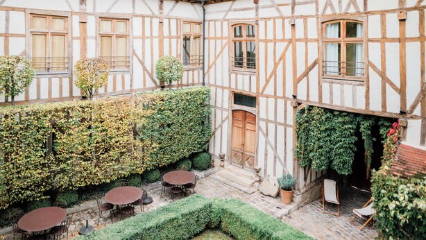 A courtyard with a traditional half-timbered building, a round table, and chairs. - La Maison de Rhodes Hôtel & Spa 5* (Troyes)