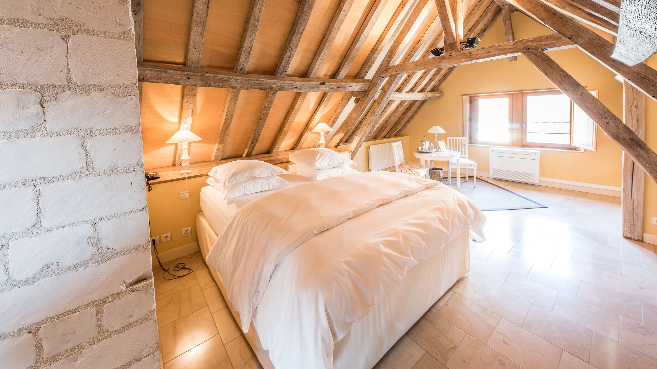 Un dormitorio con techo inclinado, vigas de madera, cama con ropa blanca, una mesita con lámpara y una ventana.