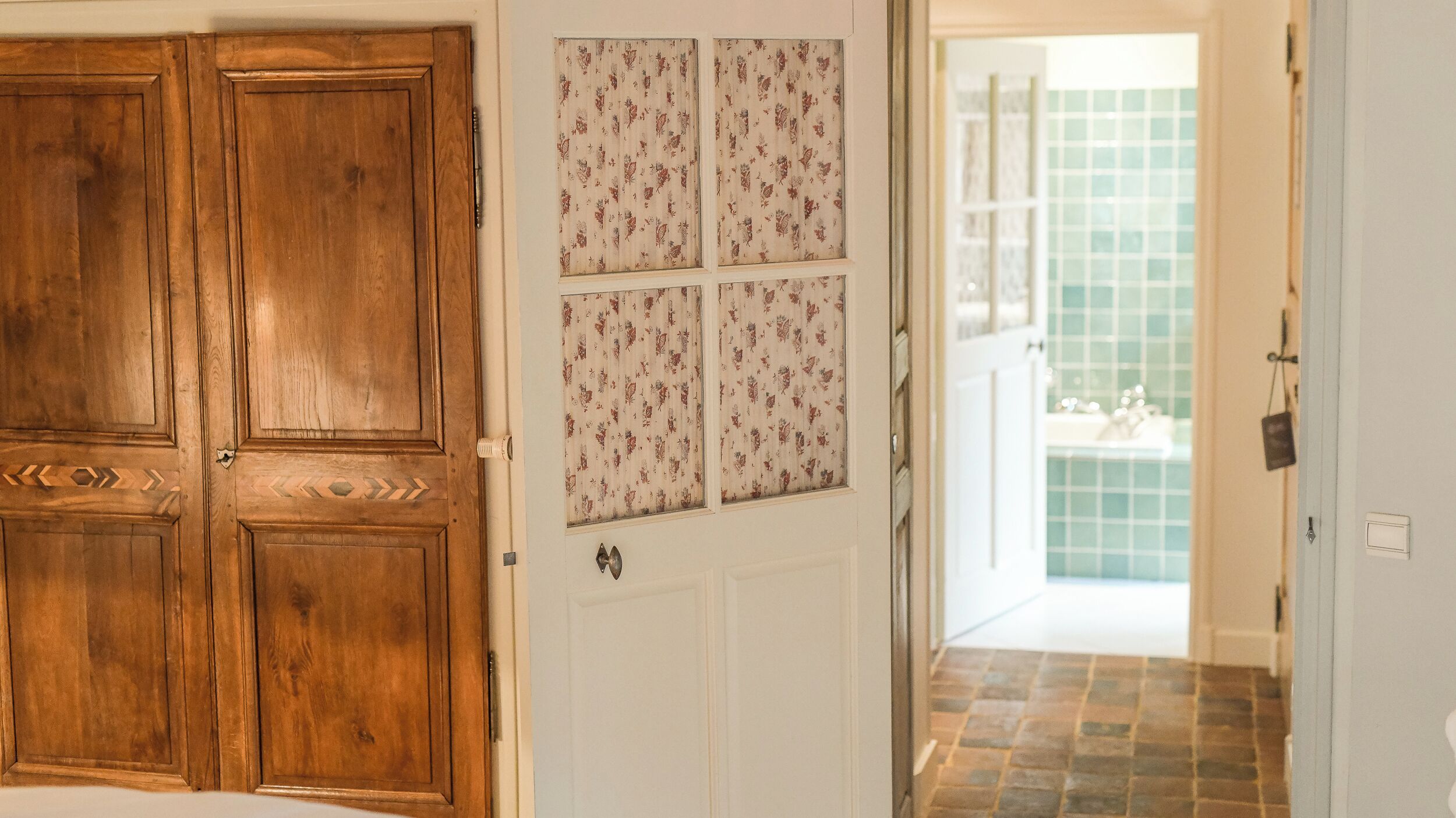 Een houten deur met een decoratief paneel, een witte deur met glaspanelen met een bloemmotief, en een uitzicht op een badkamer met een betegelde vloer en een groene wand betegeld met tegels.