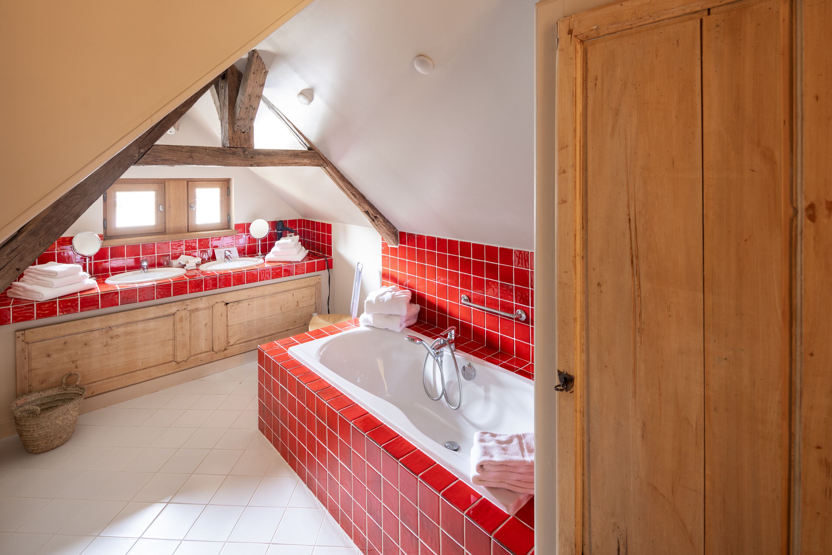 A modern bathroom with a freestanding bathtub, red tiled walls, and a wooden ceiling beam.